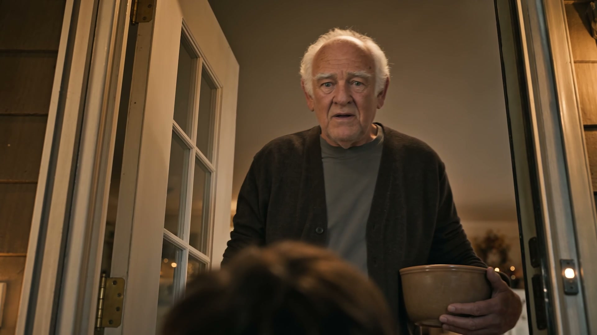 Un hombre mayor de cárdigan abre la puerta de su casa sosteniendo un bol, en luz cálida de interior, de la publicidad Cooler Kid para Wells Ferrero